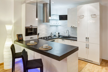 Creative interior of kitchen with furniture with neon violet light, room in beige, black and white colors. Two white cups on kitchen bar and chairs near. Elements of stones on wall.