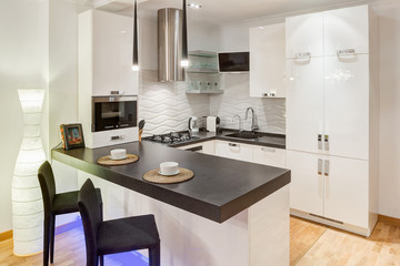 Interior of kitchen with stove, refrigerator and white furniture. Two white cups on kitchen bar, two lamp over. Kitchen in white and black colors, furniture with neon light and vase lump in corner.