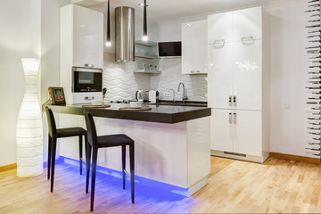 Modern interior of kitchen with white furniture, stove and refrigerator. Two white cups on kitchen bar, two lamp over. Elements of stones, white wall and black chair around.