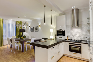 Stylish and modern interior of kitchen in white, black and green colors. Wooden table in corner of room and chair around. Two white cups on kitchen bar, lamp over.