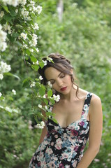 Young beautiful dark-haired woman in color dress with open shoulders in bright green blooming garden background