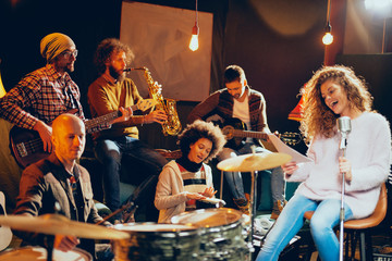 Jazz band preparing for the gig. In foreground woman singing while the rest of the band playing bass guitar, clavier and acoustic guitar. Home music studio interior.