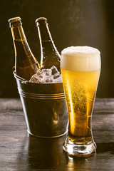 a glass of cold fresh beer with foam on the background of a bucket with ice and two-bottles of beer.