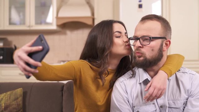 A brunette and a bearded man in glasses sitting on the corner sofa in the studio apartment; she is holding a phone and making their selfie smiling, kissing, pretending to bite his cheek, pulling faces