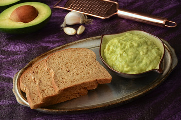 Guacamole and guacamole ingredients on purple background. Avocado, slice of bread, garlic and grater on plate