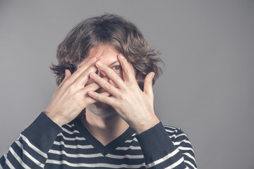 Handsome man with dark curly hair, bristle, hides face with hands, peeks through fingers, wears striped sweater, feels shy, isolated over grey background. Curious guy cannot to look at present.