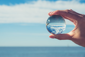 De hand van de vrouw die een kristallen bol vasthoudt, kijkt door naar de oceaan en de lucht. Creatieve fotografie, breking van kristallen bol