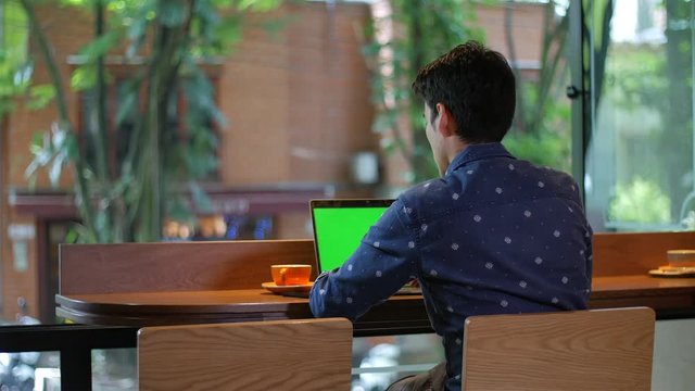 Hispanic young man sitting in coffee shop or hotel with a laptop drinking coffee, chroma key screen, medium shot. Digital nomad working or studying online. High quality footage 4K ProRes 422 HQ