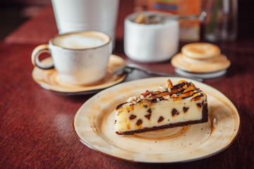 A sliced piece of cheesecake with chocolate pieces inside and poured with caramel on top. Cup with coffee in the background.
