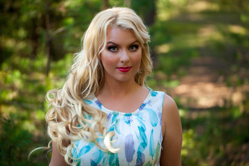 Young beautiful blonde woman in white dress with blue and green flowers posing in forest in summer