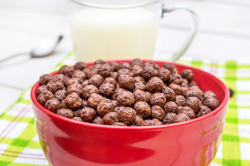 Bowl with chocolate corn balls, spoon and cup of milk on white wooden table.