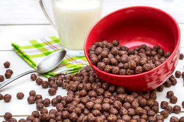 Chocolate cereal balls scattered from bowl and cup of milk on the white wooden table.
