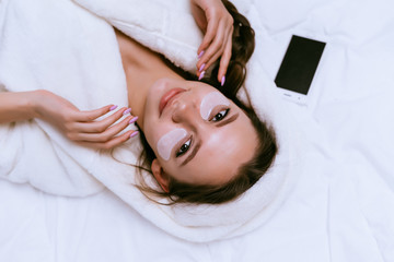 attractive young girl lies in bed, under the eyes of white silicone patches, next is a smartphone