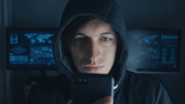 Dangerous Hacker in the hood uses a smartphone in a secret data center filled with screens
