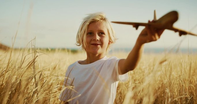 Happy young boy flying his wooden toy airplane in golden field at sunset