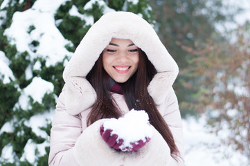 Portrait of young beautiful emotional woman in hooded down coat and gloves on snow covered garden background. Winter snowy landscape