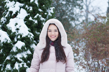 Portrait of young beautiful emotional woman in hooded down coat and gloves on snow covered garden background. Winter snowy landscape