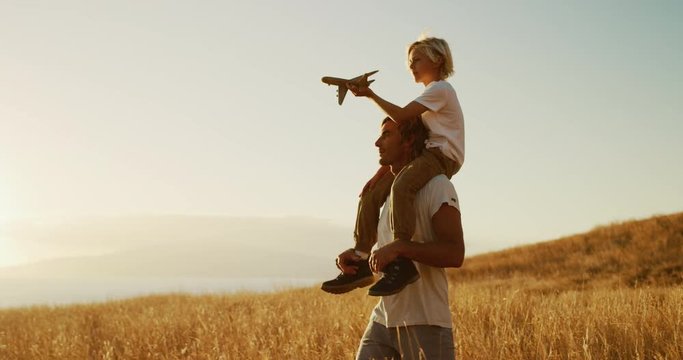 Handsome father carrying adorable son on his shoulders with toy airplane, sunset golden field