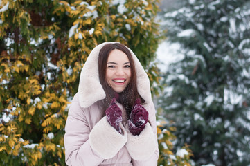 Portrait of young beautiful emotional woman in hooded down coat and gloves on snow covered garden background. Winter snowy landscape