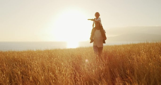 Father carrying his son on his shoulders walking through golden field at sunset