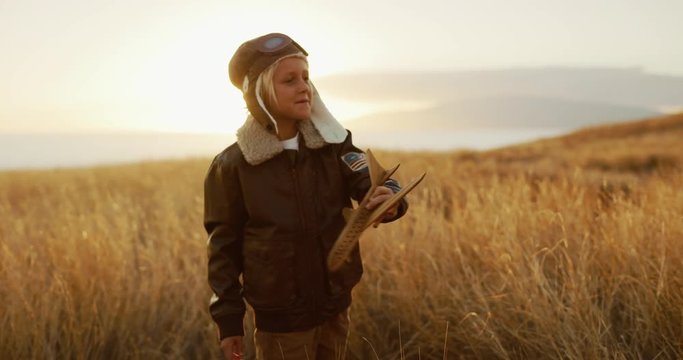 Adorable young boy wearing vintage flight suit flying his toy wooden airplane, sunset