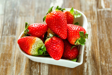 strawberries in a white plate in the shape of a heart against wooden table background