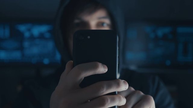 Dangerous Hacker in the hood uses a smartphone in a secret data center filled with screens