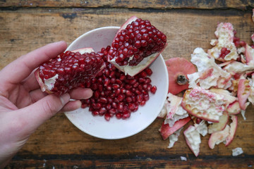 Man holds a pomegranate slice in his hand against the background of a plate with pomegranate fruits in a white plate, near the rind from the pomegranate.