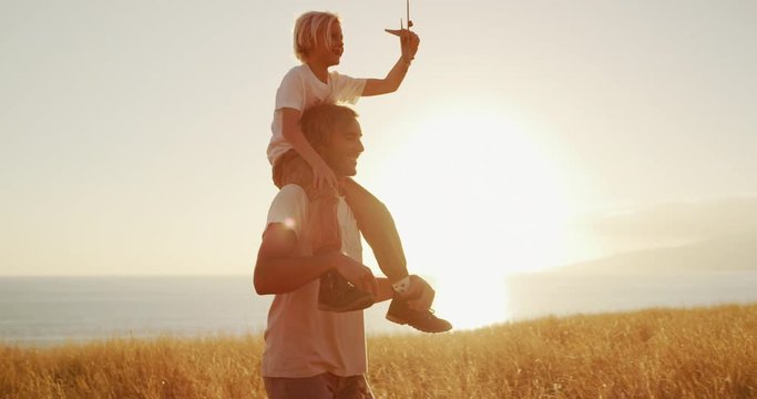Handsome father carrying adorable son on his shoulders with toy airplane, sunset golden field