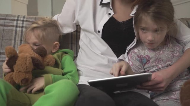Happy mother and two small kids sitting on the sofa. Woman shows tablet to children. Family holiday.