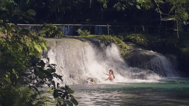Happy woman enjoying flowing water from tropical waterfall. Young woman bathing in waterfall, flowing stream water splashing on background