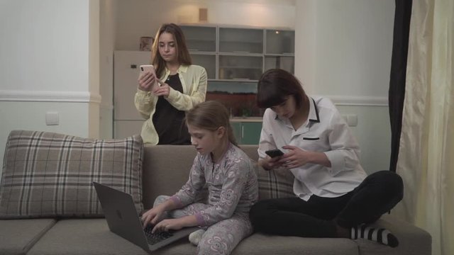 Two older sisters texting messages on cell phones younger girl typing on laptop sitting on the couch in the room. Family spends time together but not pay attention to each other.