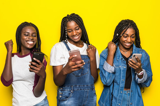 Portrait of three happy young african woman friends smiling while using mobile phone together with win gesture isolated on yellow background
