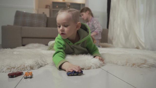 Little boy with stylish haircut plays with his toycars lying on the floor on fluffy carpet. Sister playing with a teddy bear on the background.