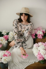 Portrait of boho girl sitting at pink and white peonies on rustic wooden floor. Stylish hipster woman in bohemian dress holding pink peony among flowers. International Womens Day