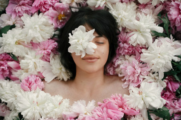 Beautiful brunette girl with white peony on eye lying in pink peonies. Aroma and care concept....
