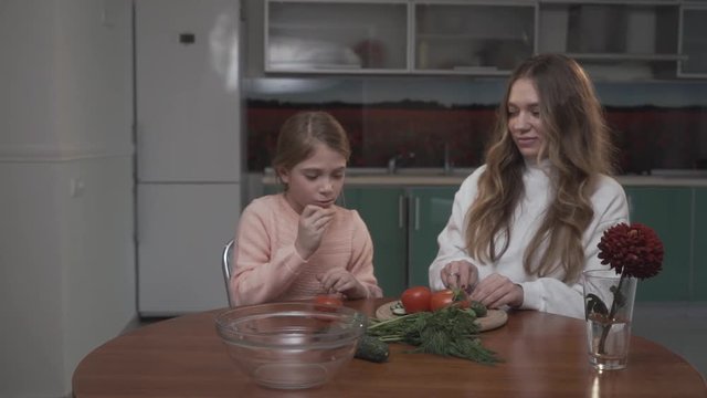 Younger sister helps the older sister coocking vegetable salad for breakfast sitting at a small table in the cozy kitchen. Sisters relationship.