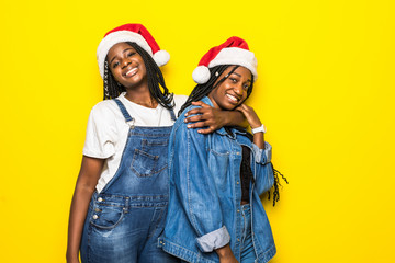 Fototapeta premium Two cheerful girls dressed in red hats celebrating Christmas together, standing isolated over yellow background