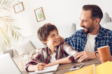 Father and son at home sitting at table dad praising boy for good grades proud while doing homework