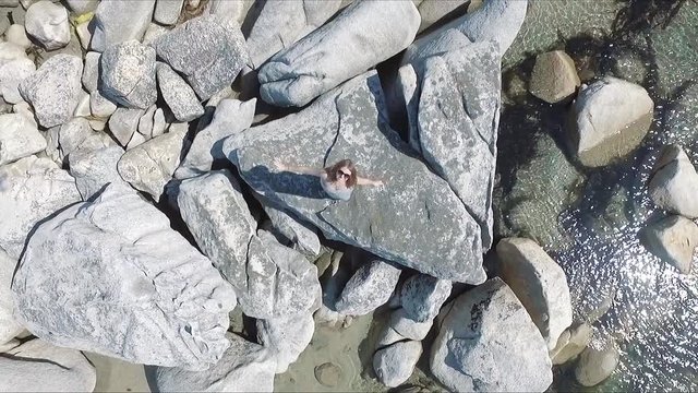 Top drone view of smiling europen girl standing on the big rock in the sea, looking to the camera and waving hands.