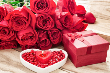 Festive background to the Valentine's day. A bouquet of red roses, a gift box, a heart-shaped plate and a heart-shaped candle. On a wooden background.