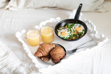 Classic breakfast. Fried eggs with vegetables in black pan, two croissants with freshly squeezed orange juice on a tray standing on white bed. Breakfast for two in bed.