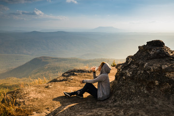A woman sitting  on rocky mountain drinking water and looking out at scenic natural view and beautiful blue sky