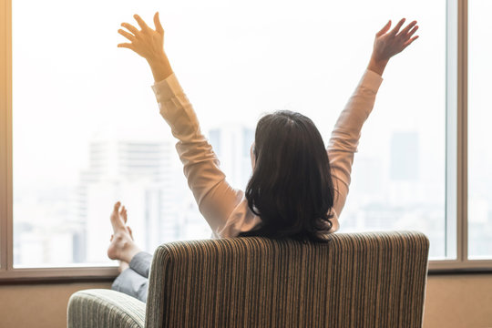 Life quality concept with business woman rear view relaxing sitting in rest on cozy armchair in modern hotel guest room or luxury home living room looking toward beautiful city urban scene