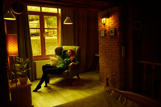 Ginger lady posing in arm chair in loft apartment in Tbilisi. She wears green shirt and black jeans. Her hair is red and curly. Walls are of red bricks. Photo has vintage style and is yellowish.