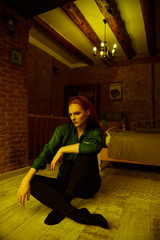 Ginger lady posing in loft apartment in Tbilisi. She wears green shirt and black jeans. Her hair is curly. Flat has big window. Walls are of red bricks. Photo has vintage style and is yellowish.