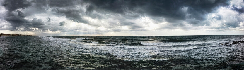 Naklejka na ściany i meble Dramatic weather panoramic view over the sea threatening waves crashing at the shore with overcast sky and sunbeams on the water - find more in my profile