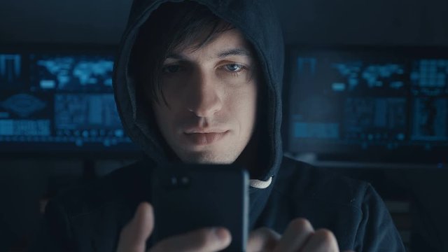 Dangerous Hacker in the hood uses a smartphone in a secret data center filled with screens
