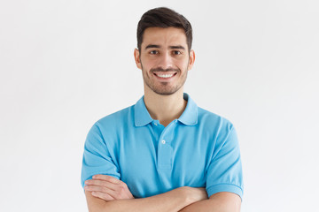 Obraz premium Uśmiechnięty przystojny mężczyzna w niebieskiej koszulce polo stojący ze skrzyżowanymi rękami na białym tle na szarym tle