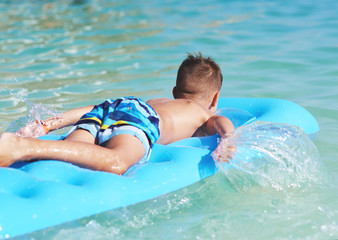 European boy is swimming in the sea on the blue air mattress, making water splashes. He is happy...